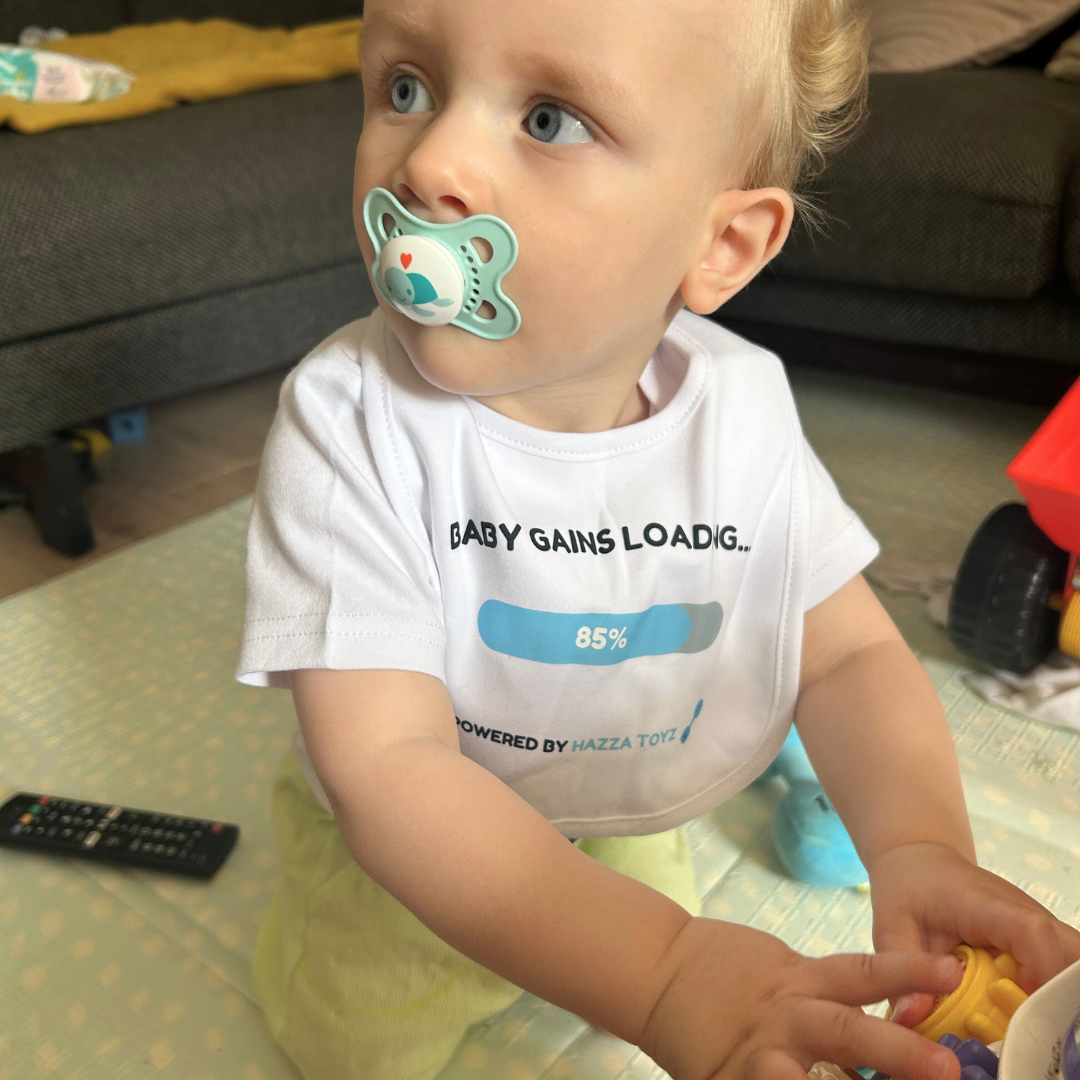 Baby wearing a “Baby Gains Loading… 85%” bib powered by Hazza Toyz, sitting on a play mat with a teal pacifier and playing with toys.