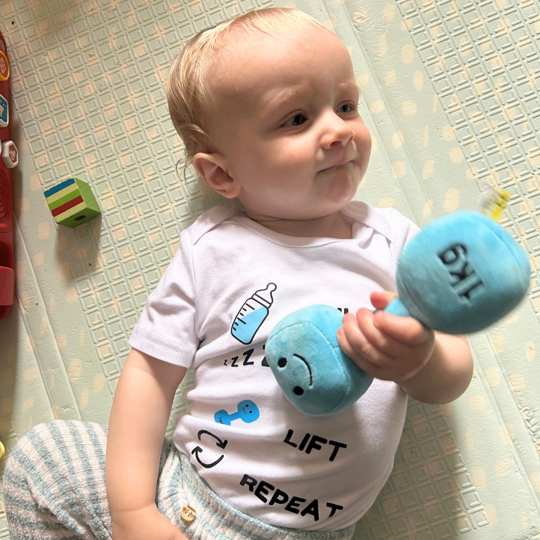 Baby wearing Milk Nap Lift Repeat bodysuit from Hazza Toyz holding blue plush dumbbell while lying on play mat