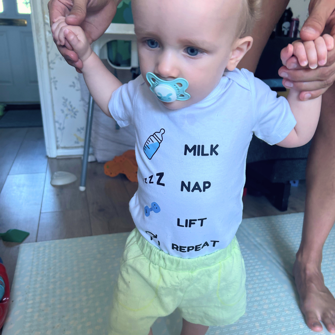 Baby wearing Milk Nap Lift Repeat bodysuit from Hazza Toyz standing with assistance and using a dummy