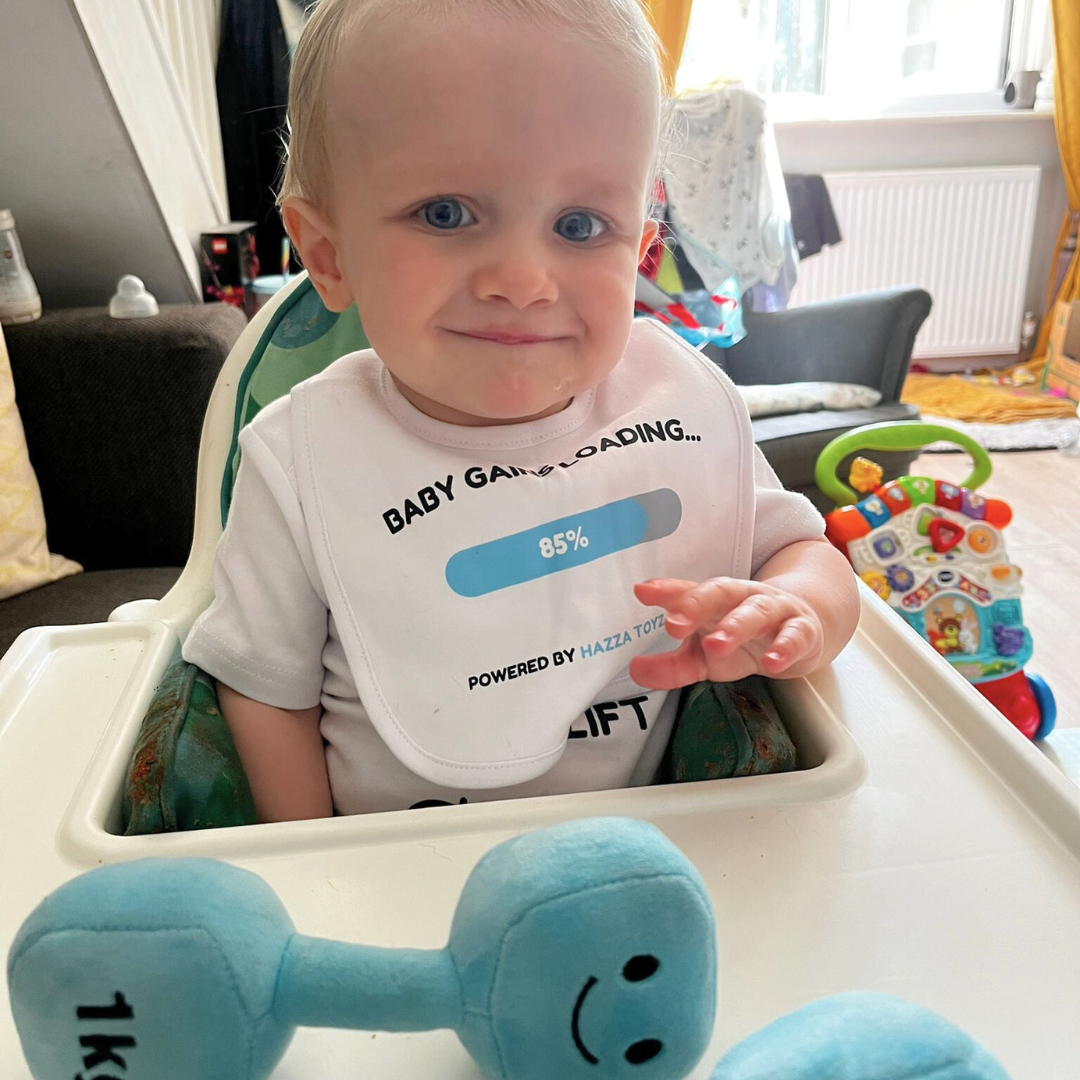 Baby wearing Baby Gains Loading gym-themed bib and bodysuit from Hazza Toyz sitting in highchair with blue plush dumbbell toy