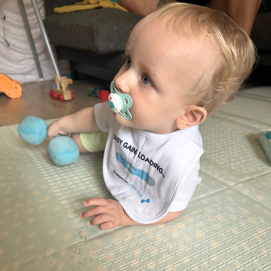 Baby wearing Baby Gains Loading bib from Hazza Toyz holding blue plush dumbbell while lying on play mat with pacifier