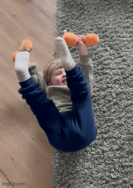 Young child lying on the floor, playfully lifting an orange plush dumbbell using both feet and one hand, smiling mid-action.