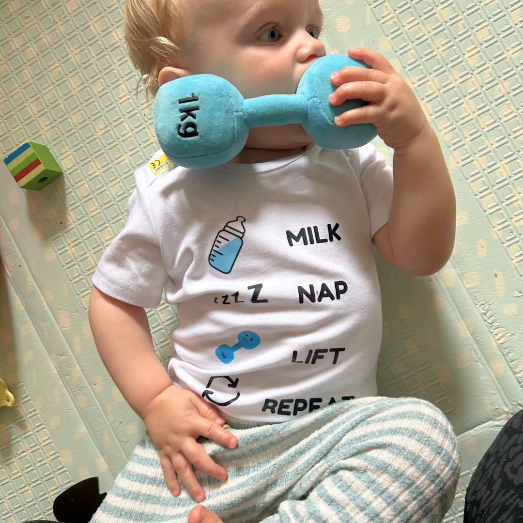Baby sitting on a playmat, holding a blue Hazza Toyz plush dumbbell near their mouth while wearing the βMilk Nap Lift Repeatβ bodysuit featuring baby bottle, nap, and dumbbell icons in Hazza Toyz blue.