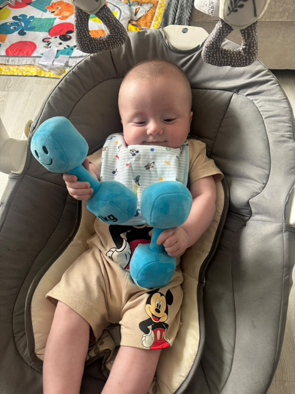 Baby relaxing in bouncer gripping Mystic Blue Hazza Toyz 1kg plush dumbbell – playful and cheeky gym-inspired baby toy for fitness-loving families