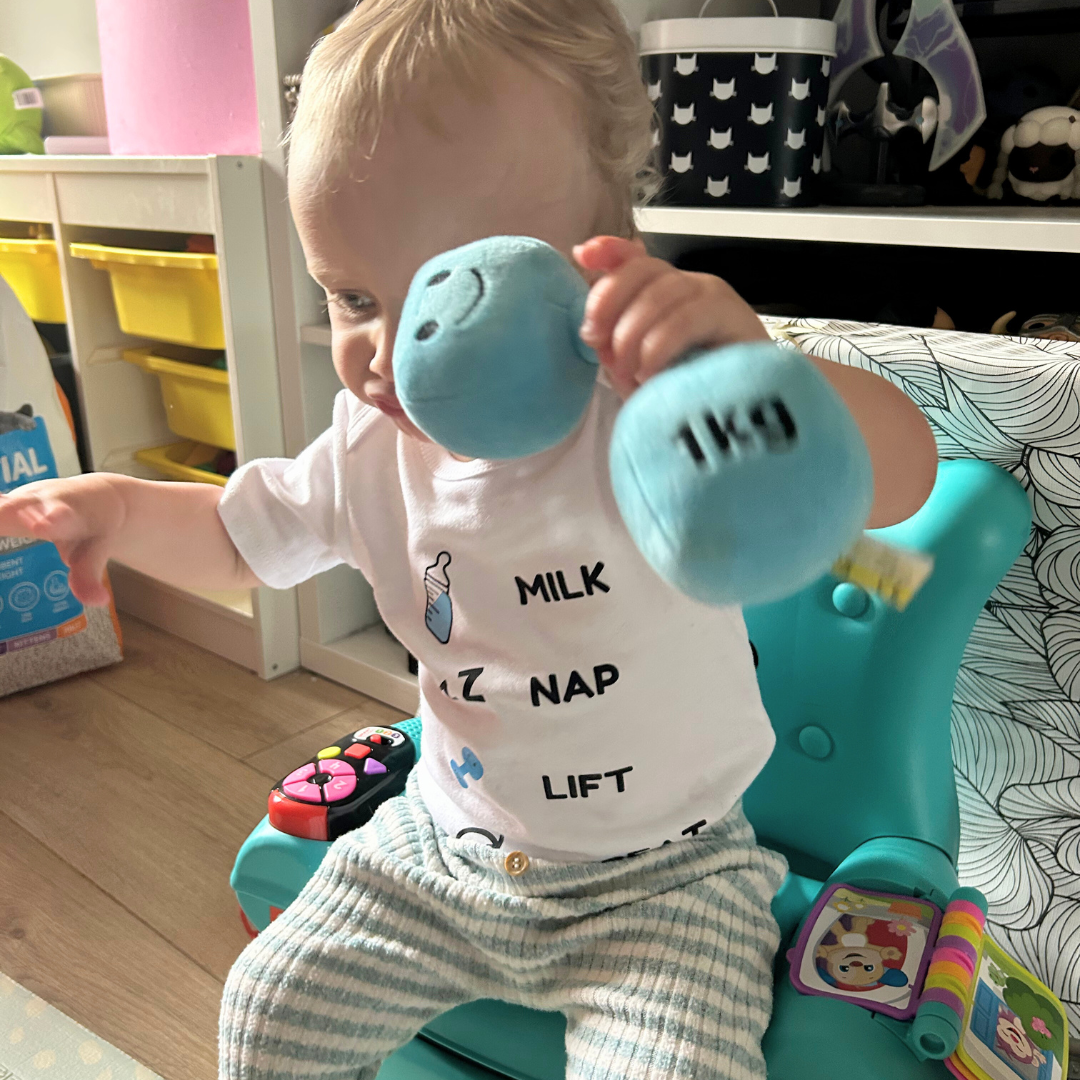 Baby sitting in a turquoise seat, holding a blue Hazza Toyz plush dumbbell while wearing the βMilk Nap Lift Repeatβ bodysuit with baby bottle, nap, and dumbbell icons.