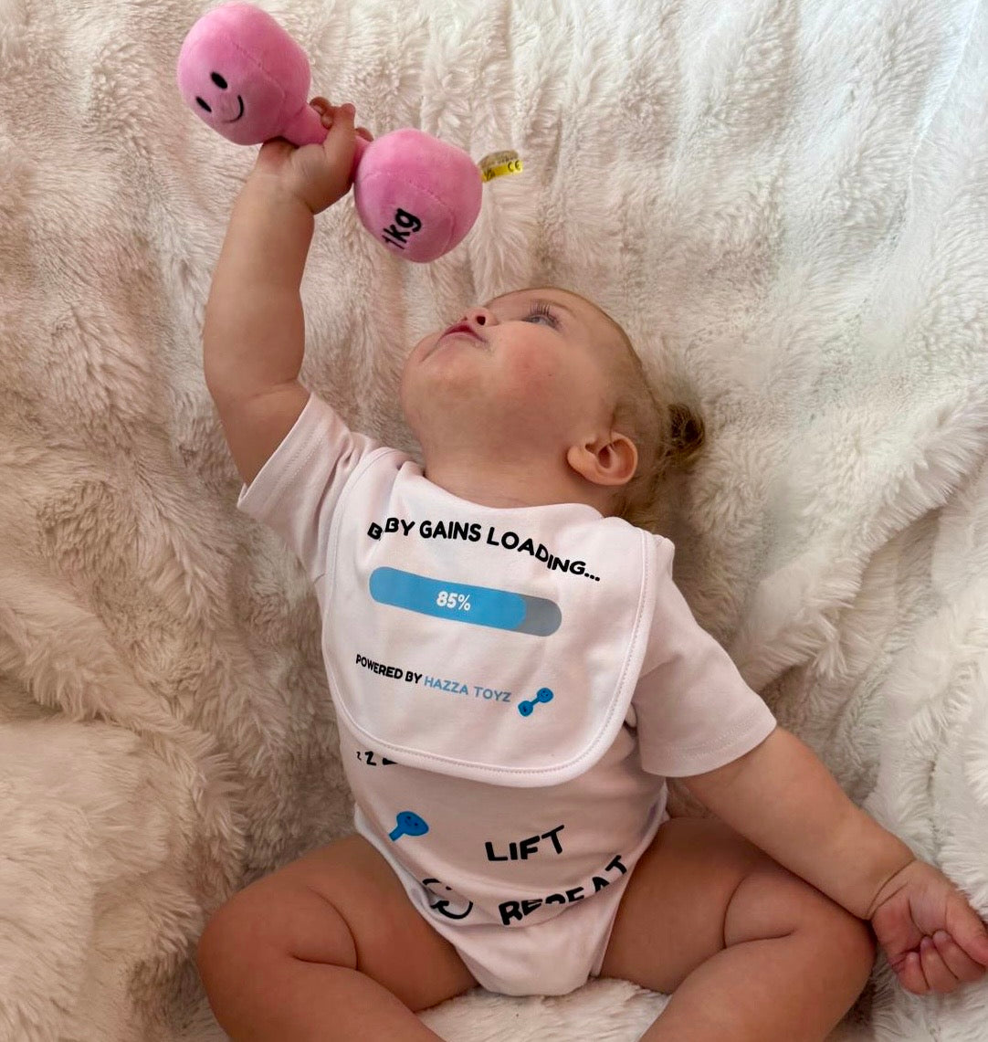Baby in Hazza Toyz bib and Milk Nap Lift Repeat bodysuit holding a pink plush 1kg dumbbell above their head, looking up with determination – cute fitness-themed baby moment