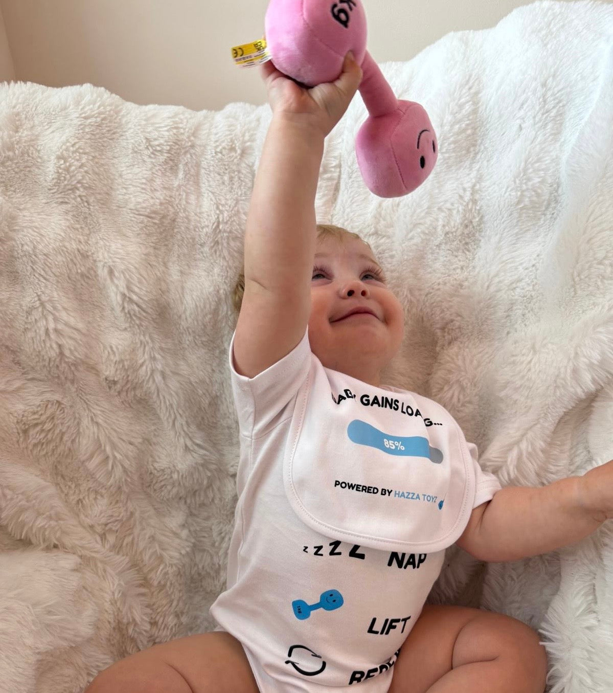 Smiling baby lifting pink 1kg plush dumbbell while wearing Hazza Toyz bib and Milk Nap Lift Repeat bodysuit – adorable fitness-themed baby photo