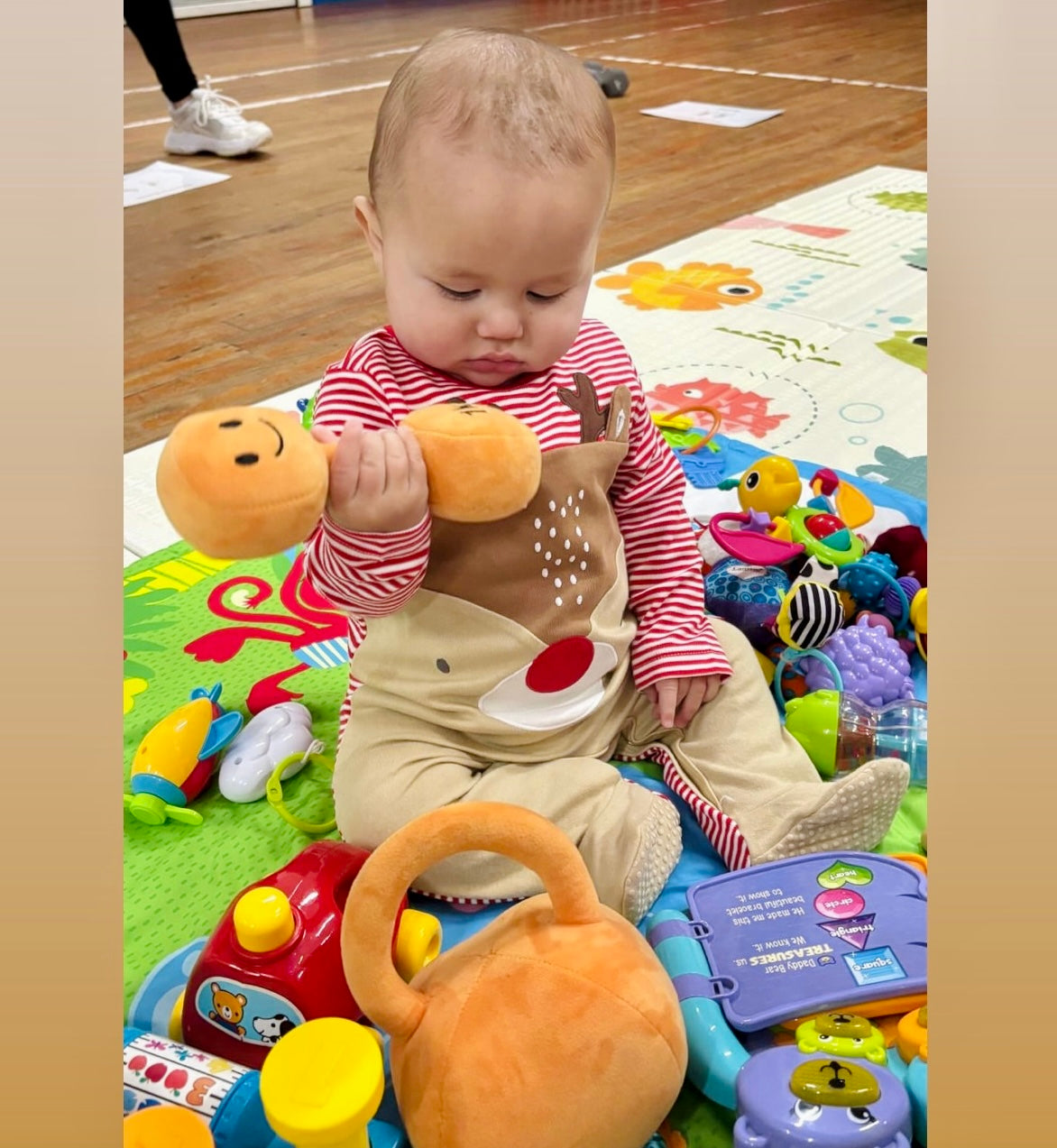 Baby sitting on colourful play mat holding Hazza Toyz plush dumbbell with matching kettlebell toy – perfect gym-themed baby gift for fitness parents