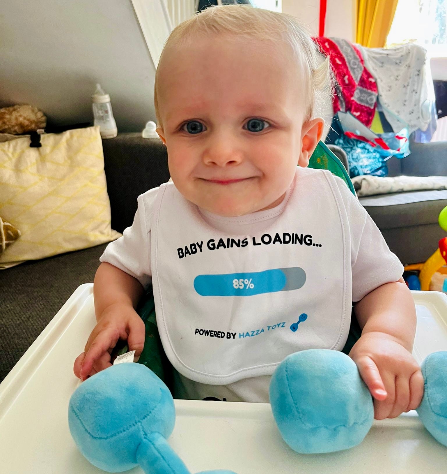 Smiling baby in high chair wearing Hazza Toyz Baby Gains Loading bib and holding Mystic Blue 1kg plush dumbbell – funny gym-themed baby gift for fitness-loving parents