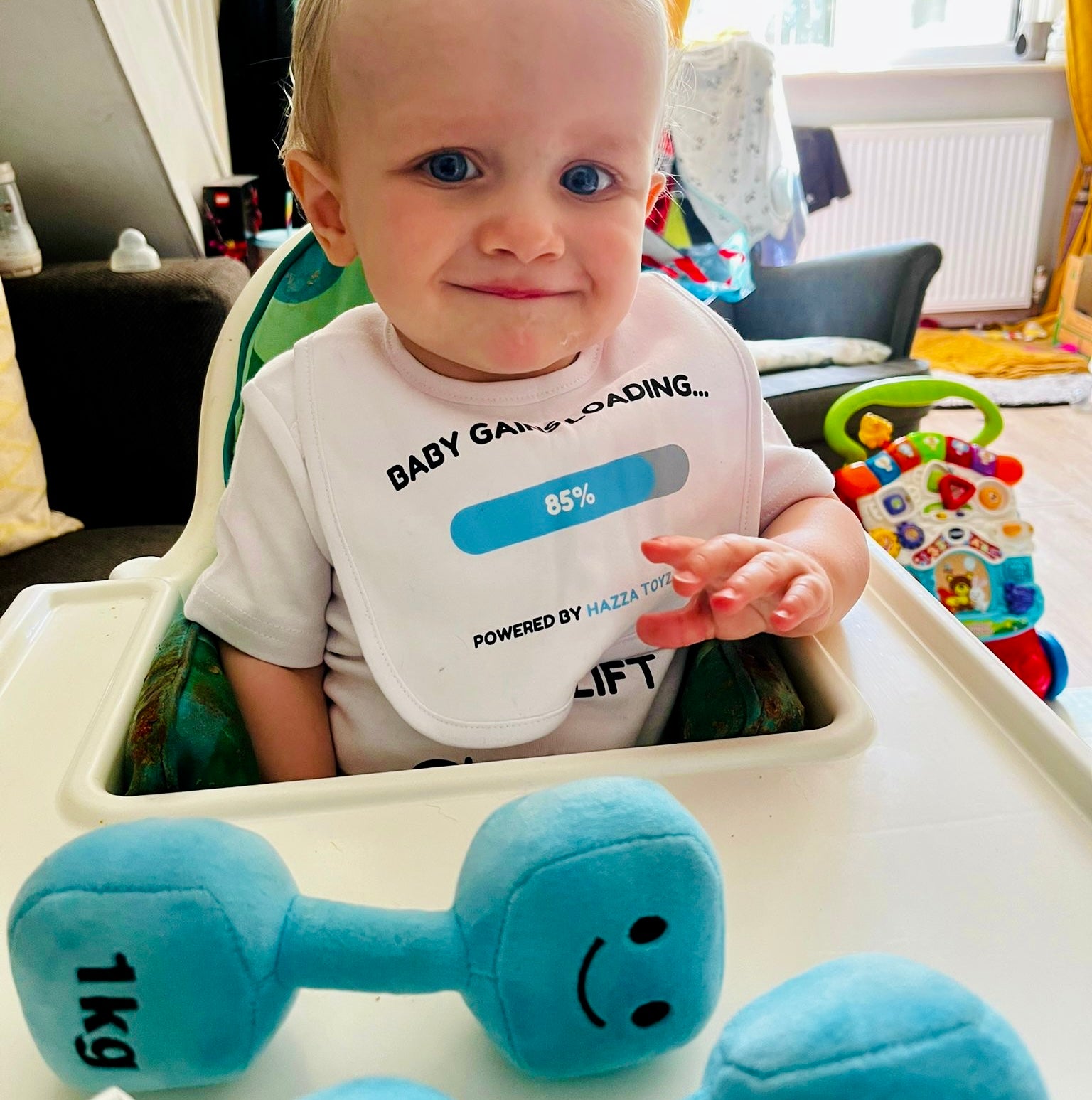 Baby sitting in high chair wearing Hazza Toyz Baby Gains Loading bib, smiling and holding Mystic Blue 1kg plush dumbbell – cheeky gym-themed baby gift for fitness parents