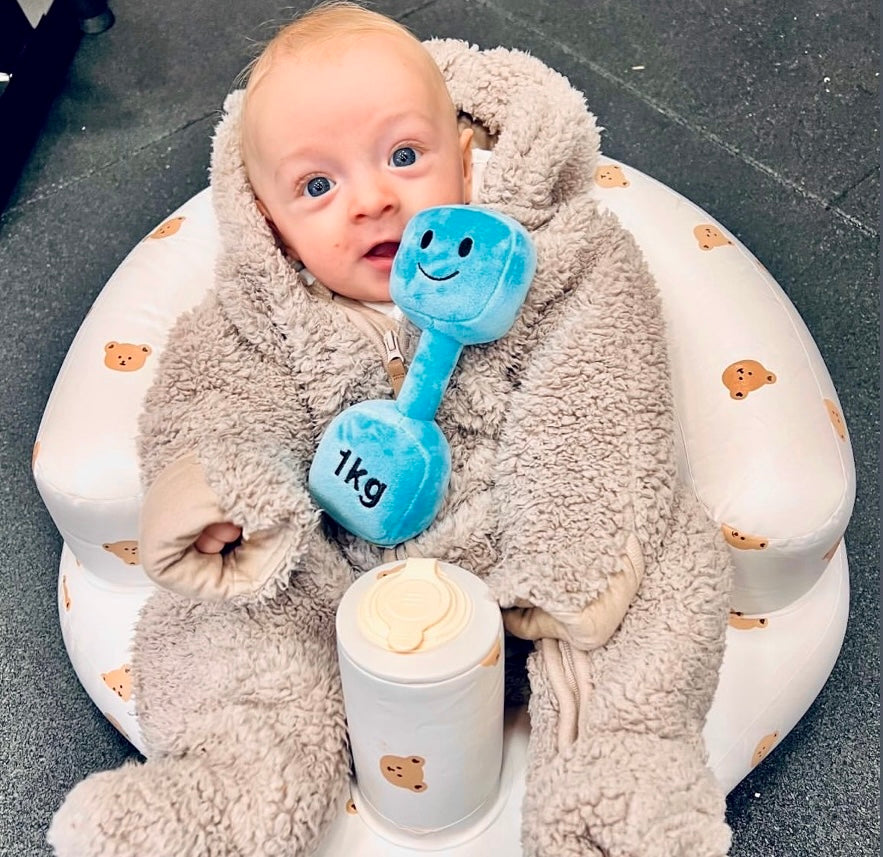 Baby sitting snug in bear-print cushion wearing a fluffy onesie, holding Mystic Blue Hazza Toyz 1kg plush dumbbell – adorable fitness-themed baby gift idea