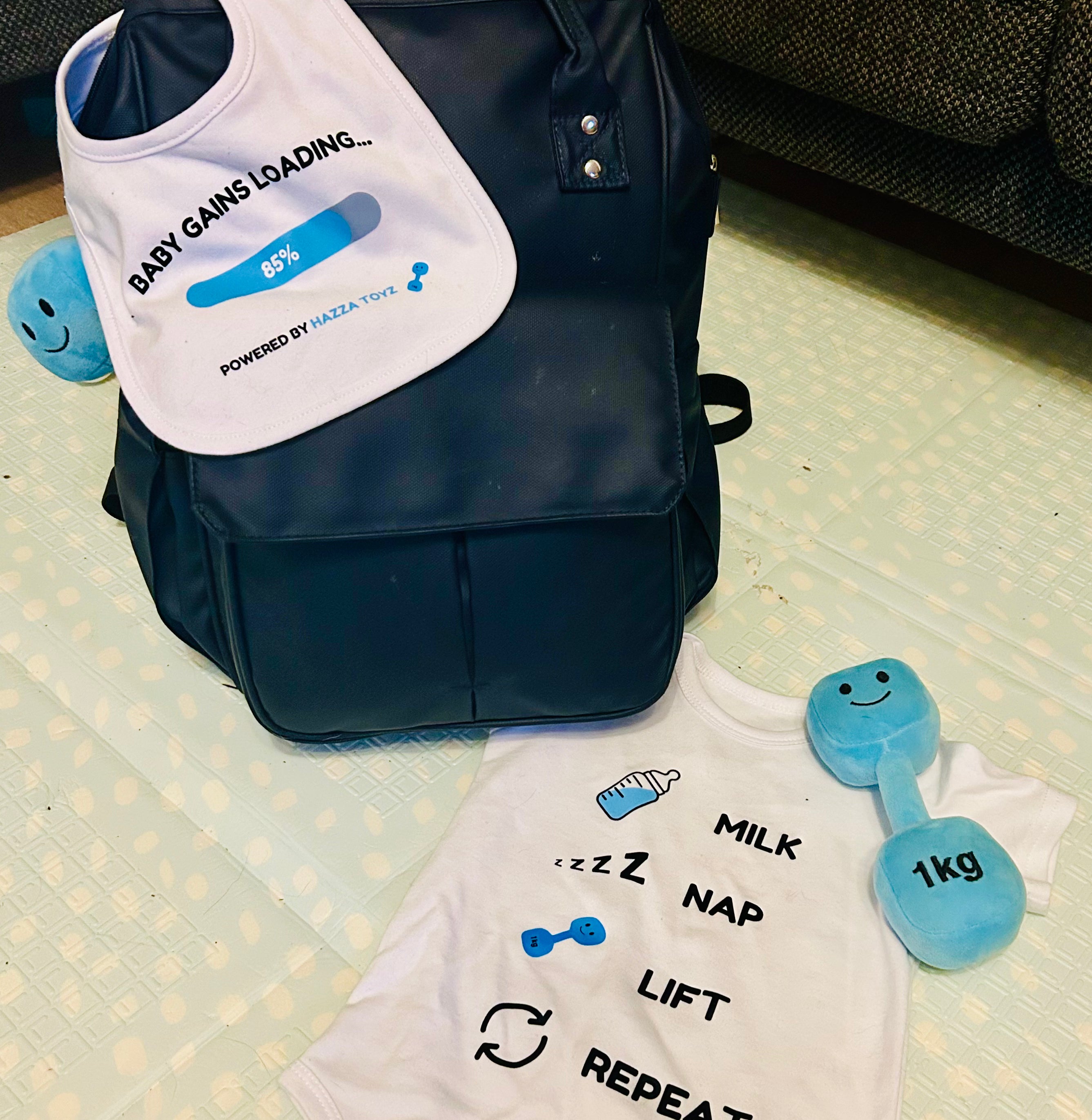 Hazza Toyz Baby Gains Loading bib draped over black changing bag, with Milk Nap Lift Repeat baby bodysuit and Mystic Blue 1kg plush dumbbell – fun gym-themed baby gift set for fitness-loving parents