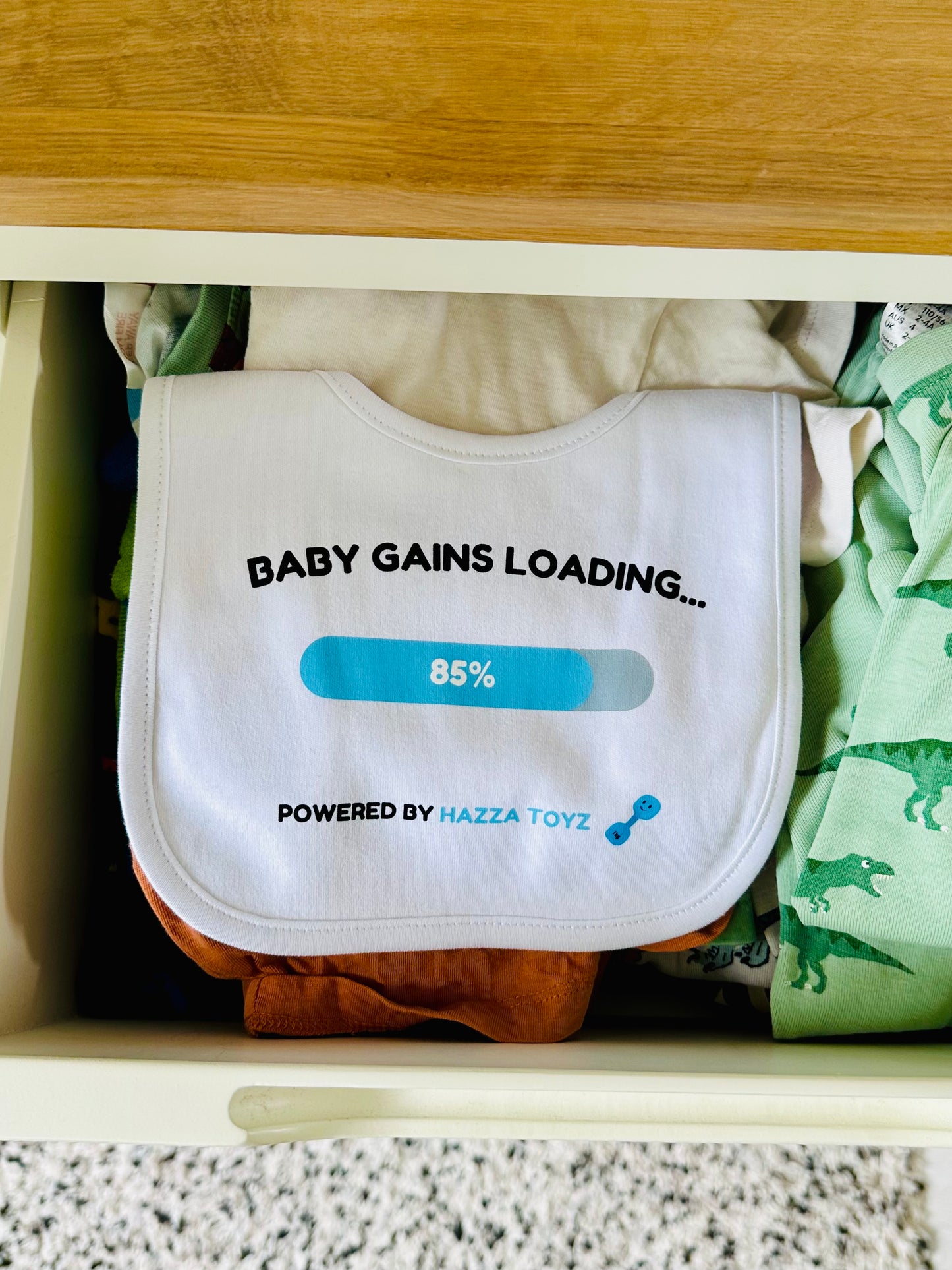 White Hazza Toyz Baby Gains Loading bib with blue progress bar and dumbbell icon, neatly placed in drawer with baby clothes – fun gym-inspired gift for newborns