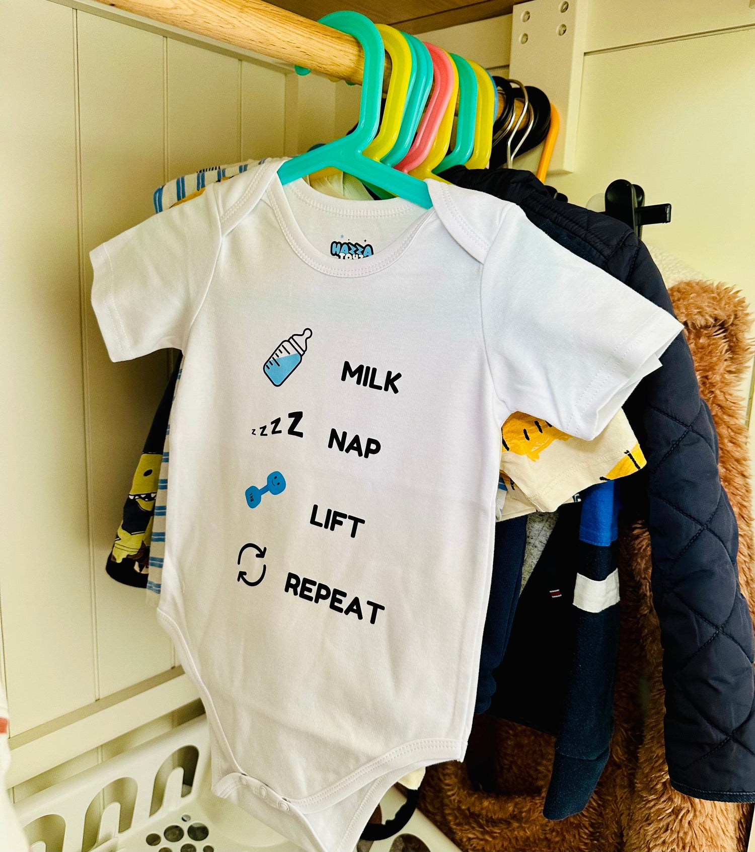 White Hazza Toyz Milk Nap Lift Repeat baby bodysuit with bottle, sleep, dumbbell, and repeat icons hanging on colourful hanger in baby’s wardrobe – fun gym-themed baby clothing