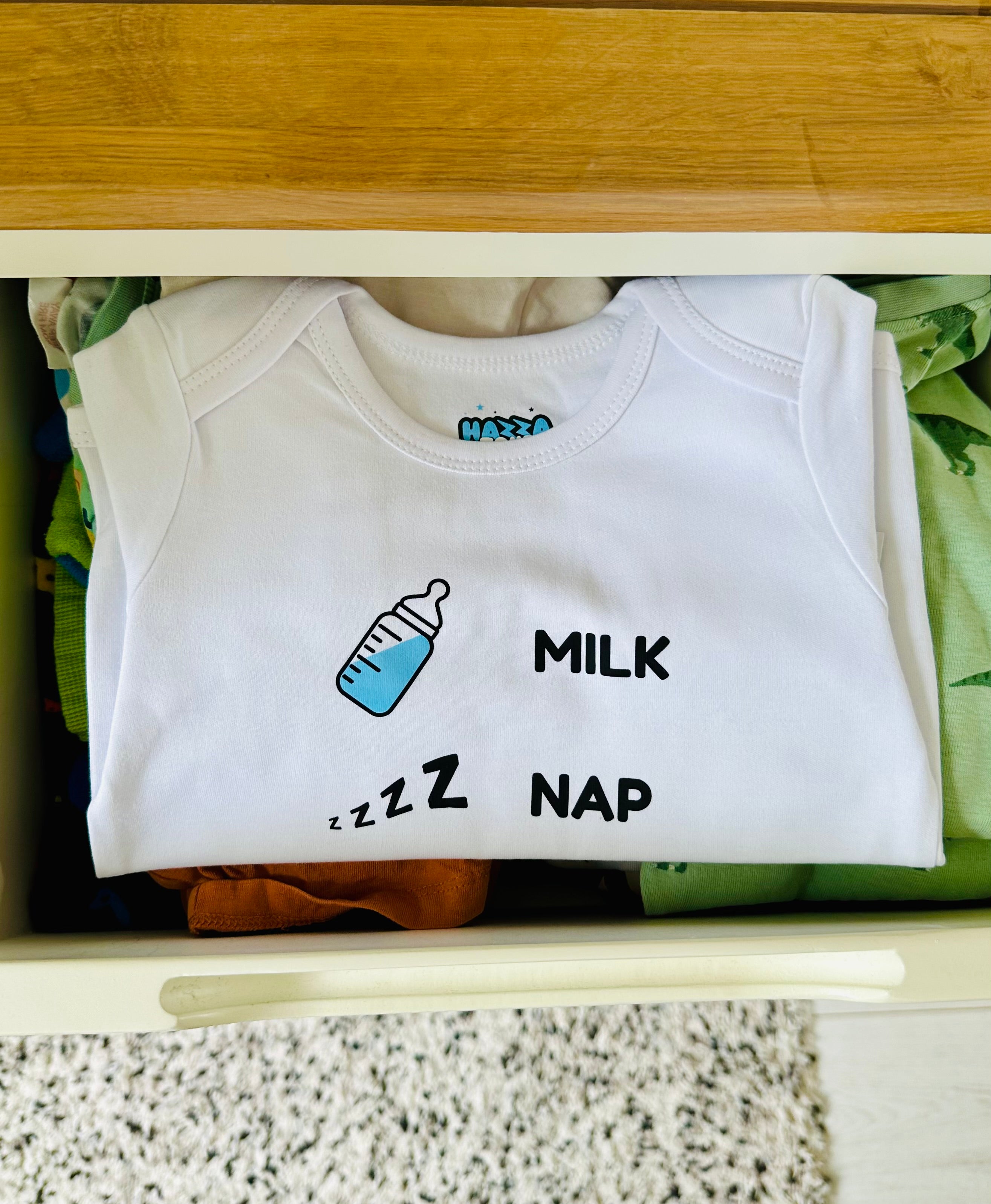 White Hazza Toyz Milk Nap baby bodysuit with blue bottle and sleep icons neatly folded in drawer – cute newborn clothing gift