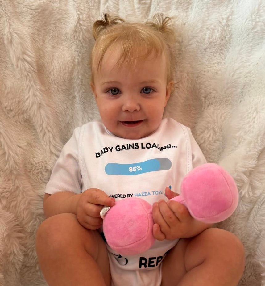 Baby sitting on a soft white blanket wearing a “Baby Gains Loading… 85%” bib, holding a pink plush dumbbell toy from Hazza Toyz.