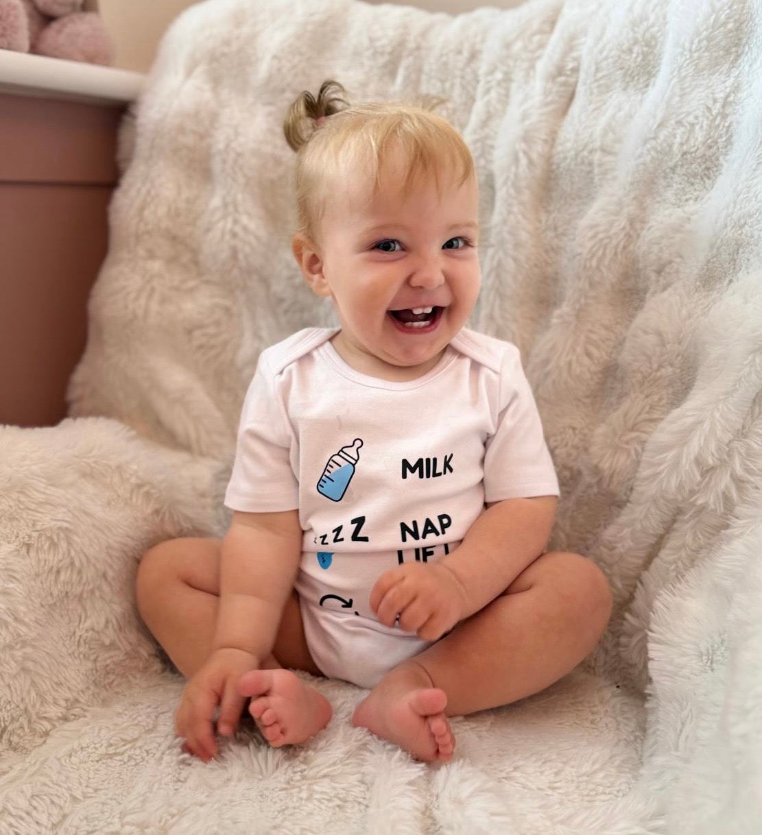 Smiling baby sitting on a fluffy white blanket wearing the Hazza Toyz βMilk Nap Lift Repeatβ bodysuit, featuring a blue baby bottle, zzz nap icon, and dumbbell graphic in Hazza Blue.