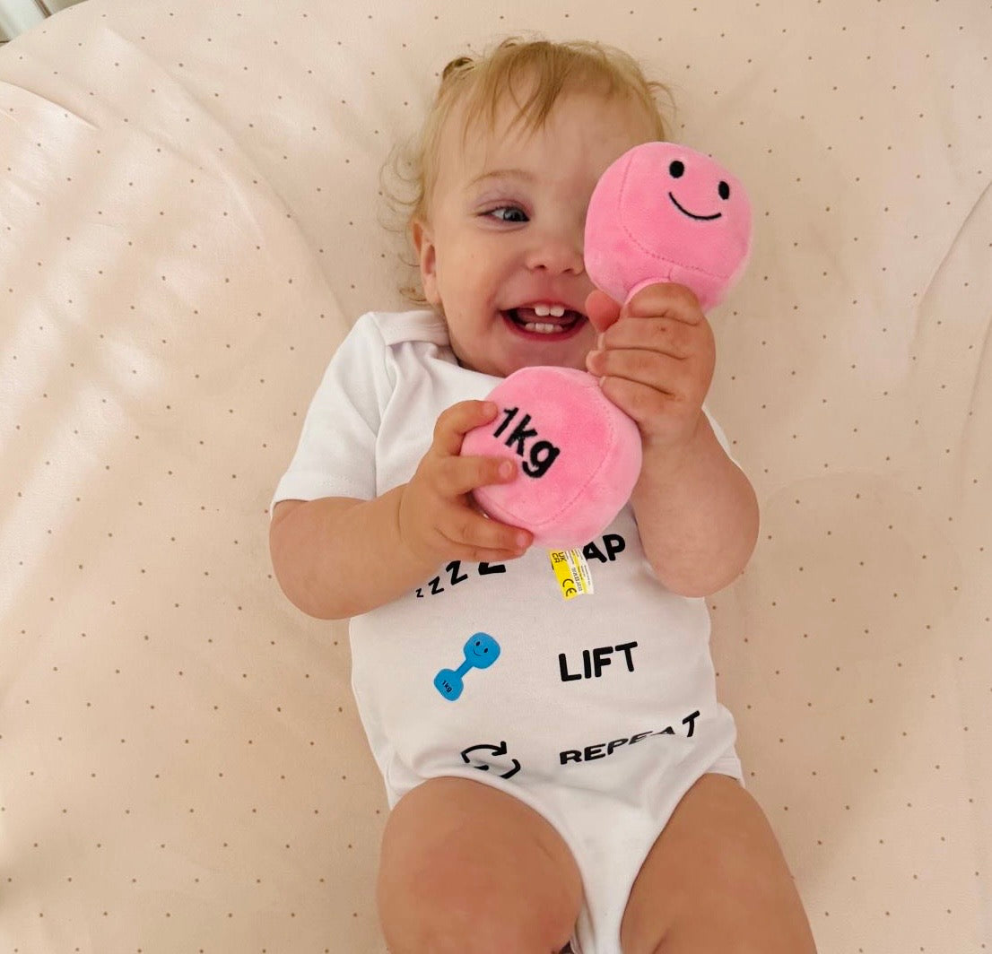 Happy baby lying on soft blanket wearing Hazza Toyz Milk Nap Lift Repeat white bodysuit and holding Candy Château Pink 1kg plush dumbbell – cute gym-themed baby gift