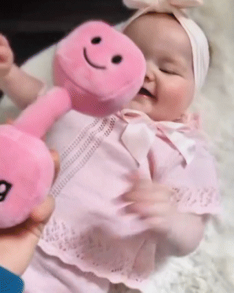 Little girl in floral romper lifting Hazza Toyz Candy Château Pink plush dumbbell and kettlebell toys – fun gym-themed baby gift in motion