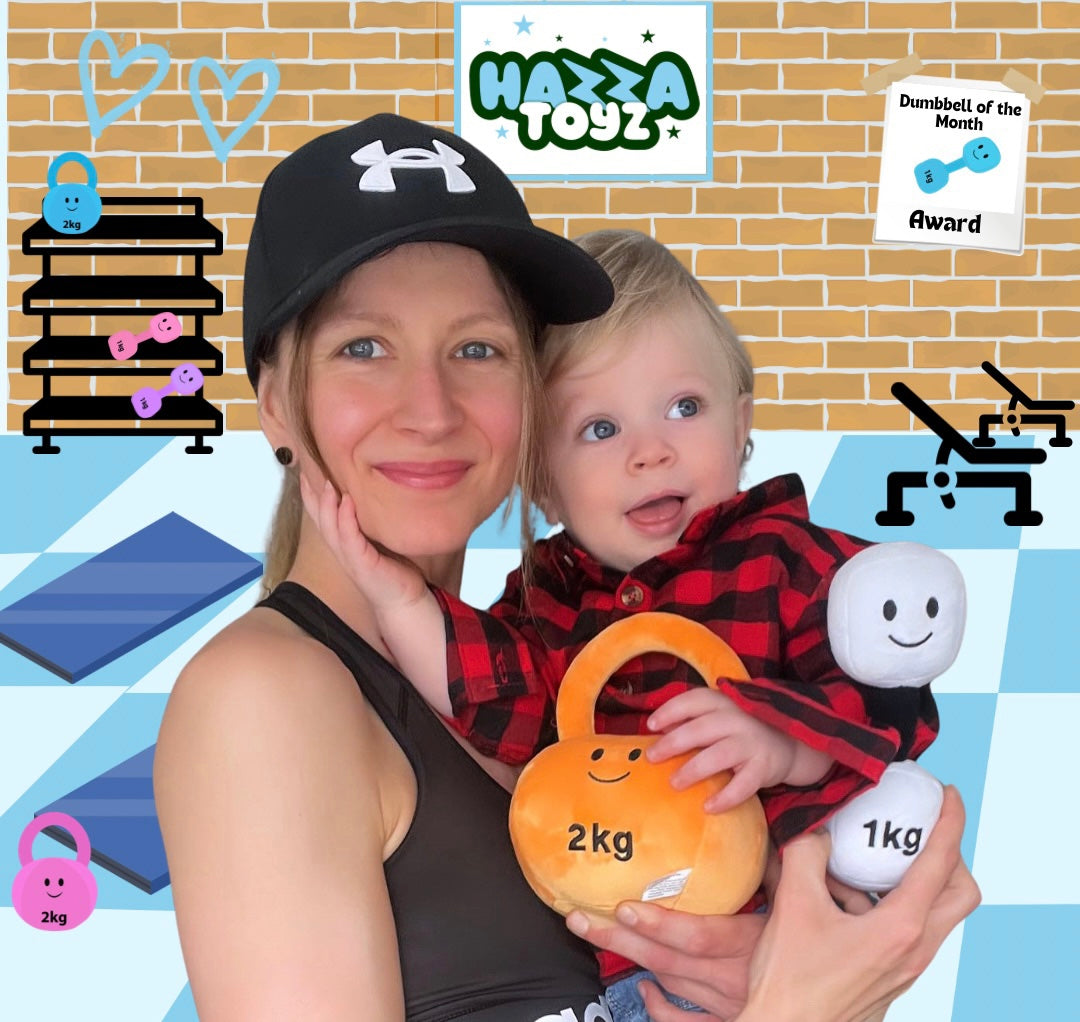 Mother holding baby in cartoon gym background with Hazza Toyz logo, as baby holds orange plush kettlebell and white plush dumbbell.