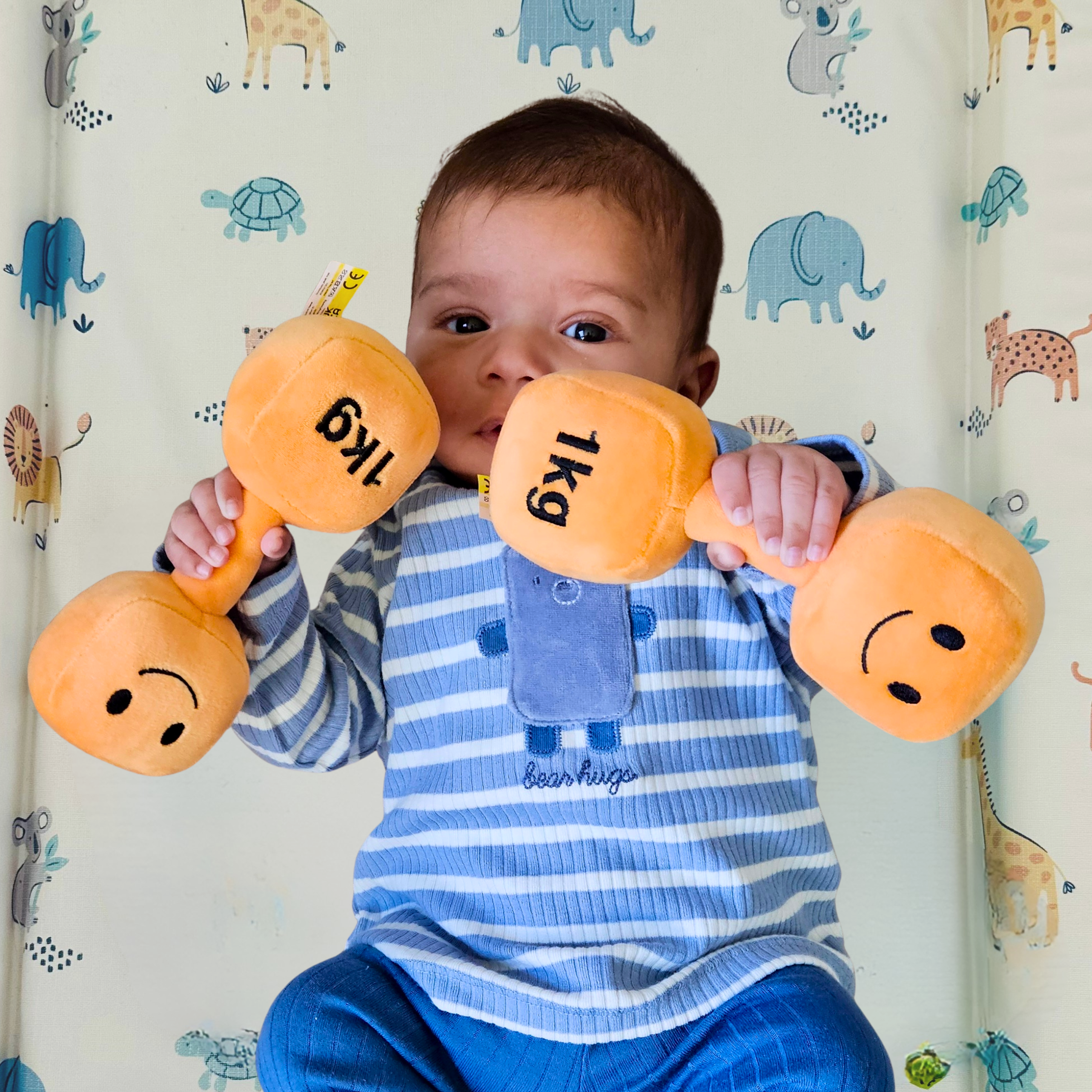Baby holding two orange plush dumbbell toys – soft 1kg sensory rattle for newborns and infants, fun fitness-inspired baby toy.