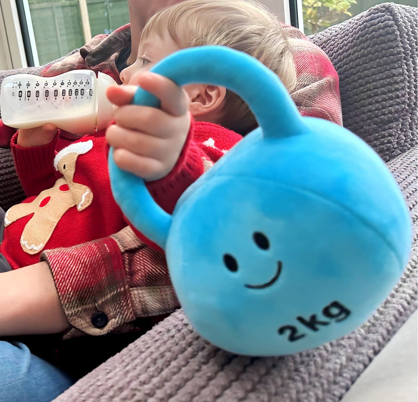 Baby holding a Mystic Blue kettlebell while drinking milk by the Christmas tree, enjoying festive playtime with Hazza Toyz fitness-inspired toys for toddlers.
