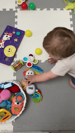 Baby reaching for and picking up a soft plush dumbbell toy – fun sensory rattle for newborn play and development.
