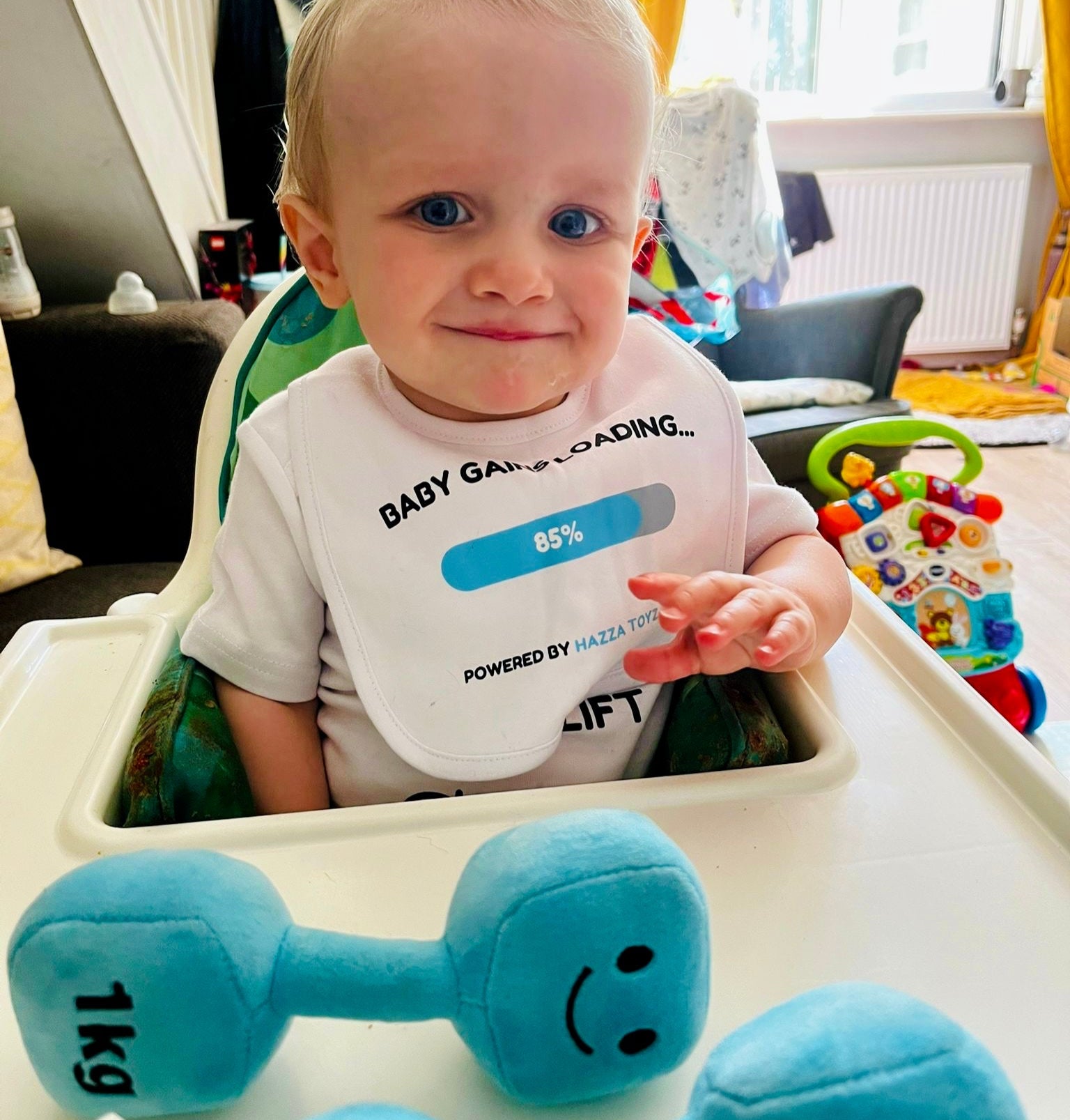 Child in a high chair with blue plush dumbbells and a shirt with text.
