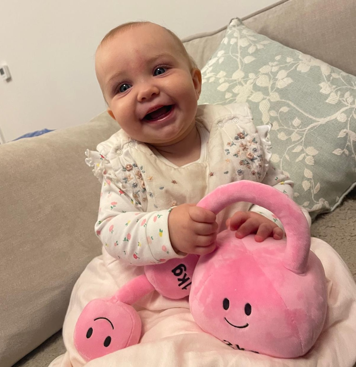 Smiling baby sitting on sofa holding pink plush kettlebell and dumbbell set