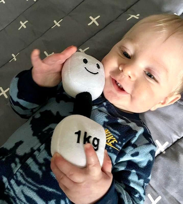 Smiling baby lying on grey blanket holding white plush dumbbell with black handle