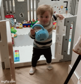 Smiling toddler walking indoors, holding a mystic blue plush kettlebell with a smiling face design, inside a play area with colourful toys in the background.