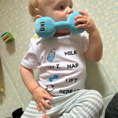 Baby sitting on a playmat, holding a blue Hazza Toyz plush dumbbell near their mouth while wearing the “Milk Nap Lift Repeat” bodysuit featuring baby bottle, nap, and dumbbell icons in Hazza Toyz blue.