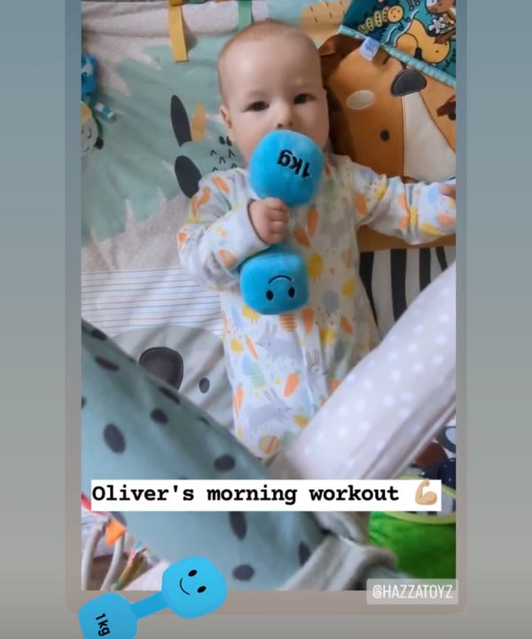 Baby in colourful sleepsuit lying on play mat and lifting Mystic Blue Hazza Toyz plush dumbbell – cheeky fitness-themed baby toy for active families
