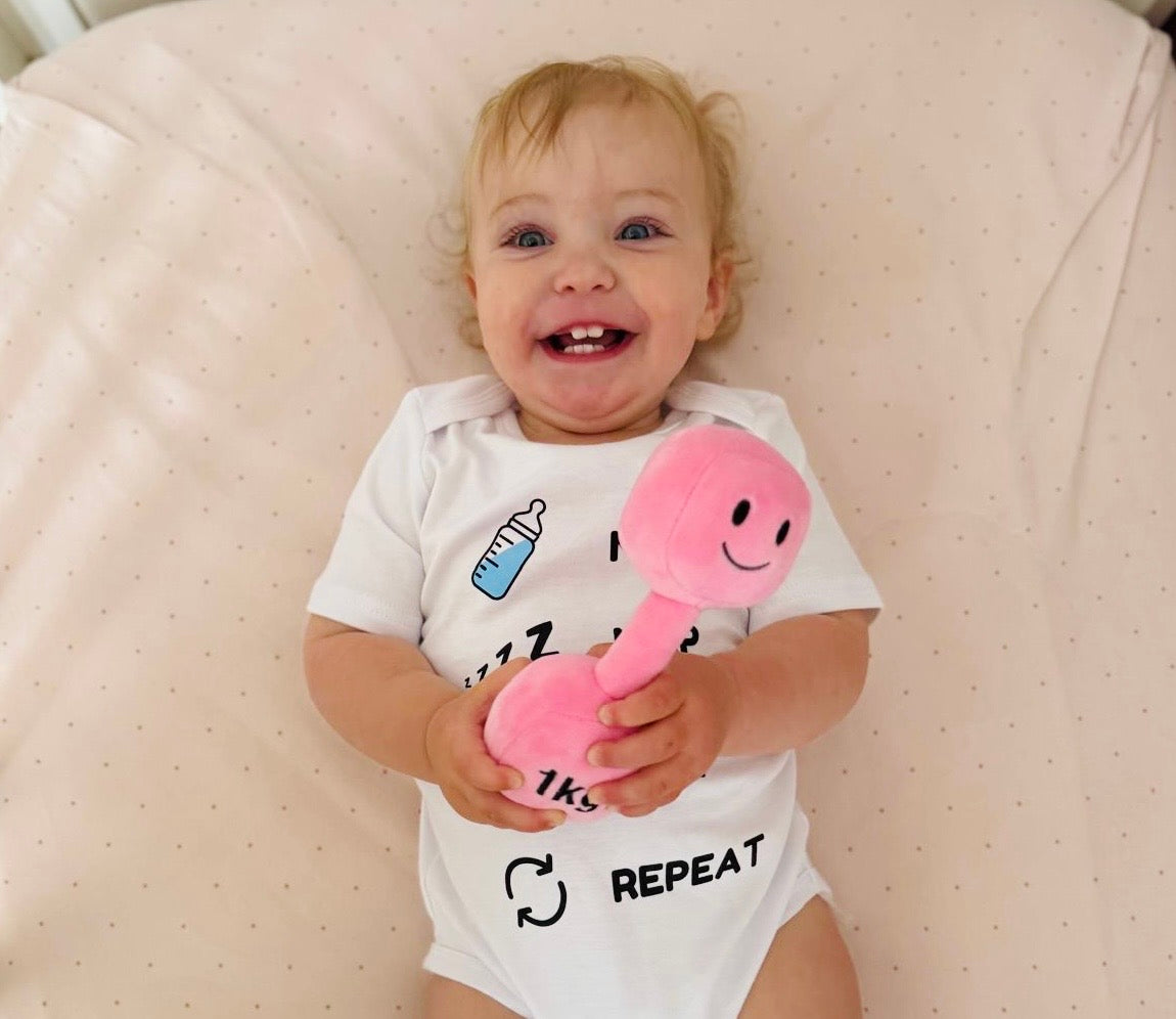 Smiling baby lying on pale pink blanket wearing Hazza Toyz Milk Nap Lift Repeat white bodysuit and holding Candy Château Pink 1kg plush dumbbell – fun gym-themed baby gift for fitness-loving parents
