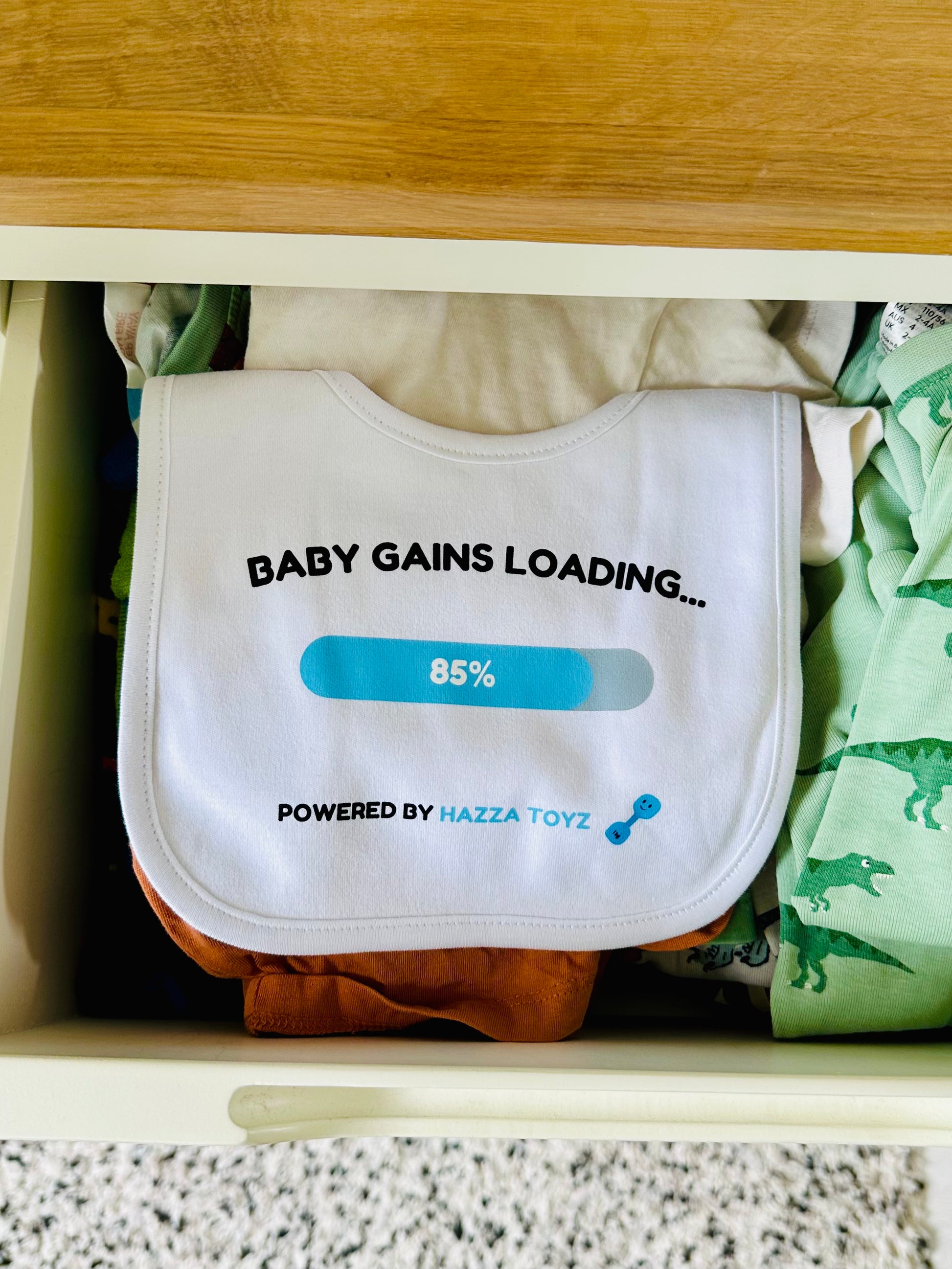 White Hazza Toyz Baby Gains Loading bib with blue progress bar and dumbbell icon, neatly placed in drawer with baby clothes – fun gym-inspired gift for newborns
