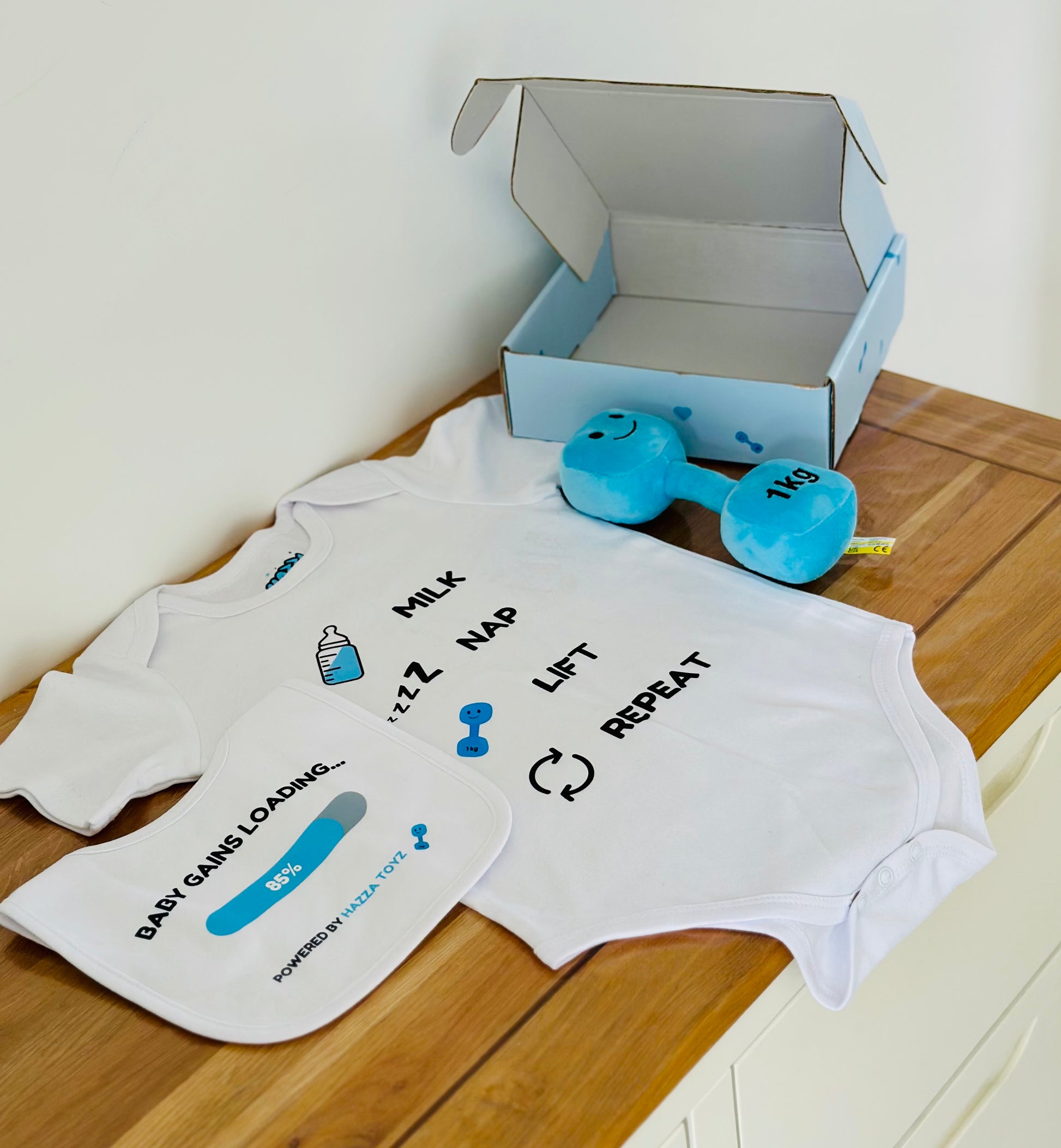 Baby onesie with text and graphics on a wooden surface next to a blue toy dumbbell and open box.