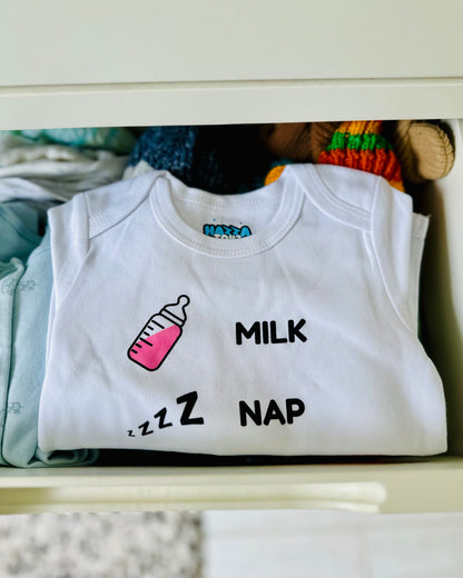 White baby bodysuit with bottle, “Milk Nap Lift Repeat” design, neatly folded in a drawer with other baby clothes.
