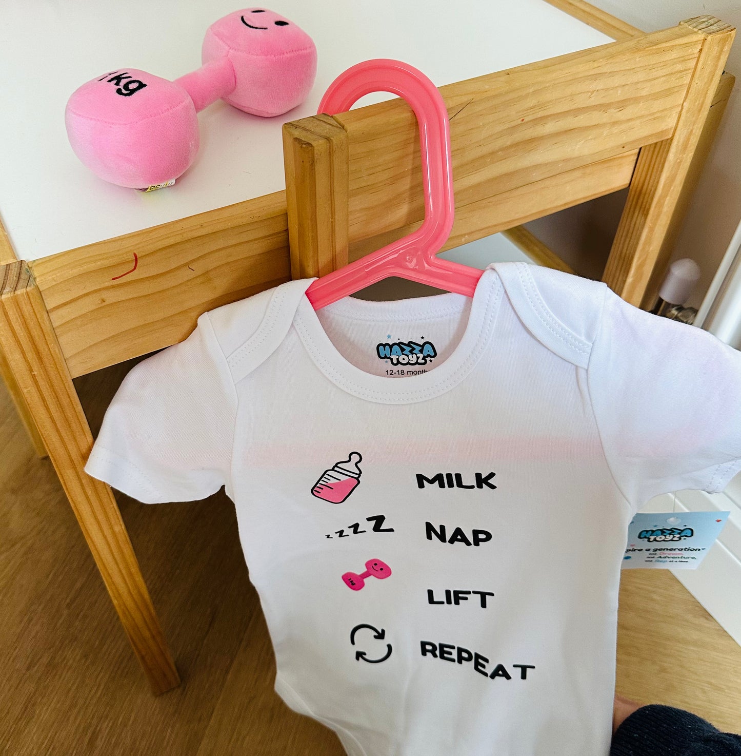 Funny baby bodysuit with “Milk Nap Lift Repeat” design on pink hanger, next to plush pink dumbbell toy on wooden table.
