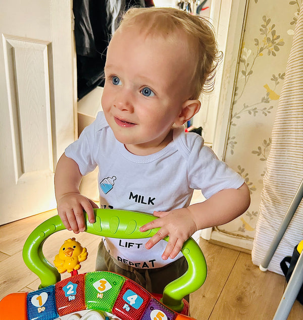Smiling toddler standing with toy walker wearing Hazza Toyz white Milk Lift Repeat baby bodysuit – fun and cheeky gym baby outfit for fitness-loving parents
