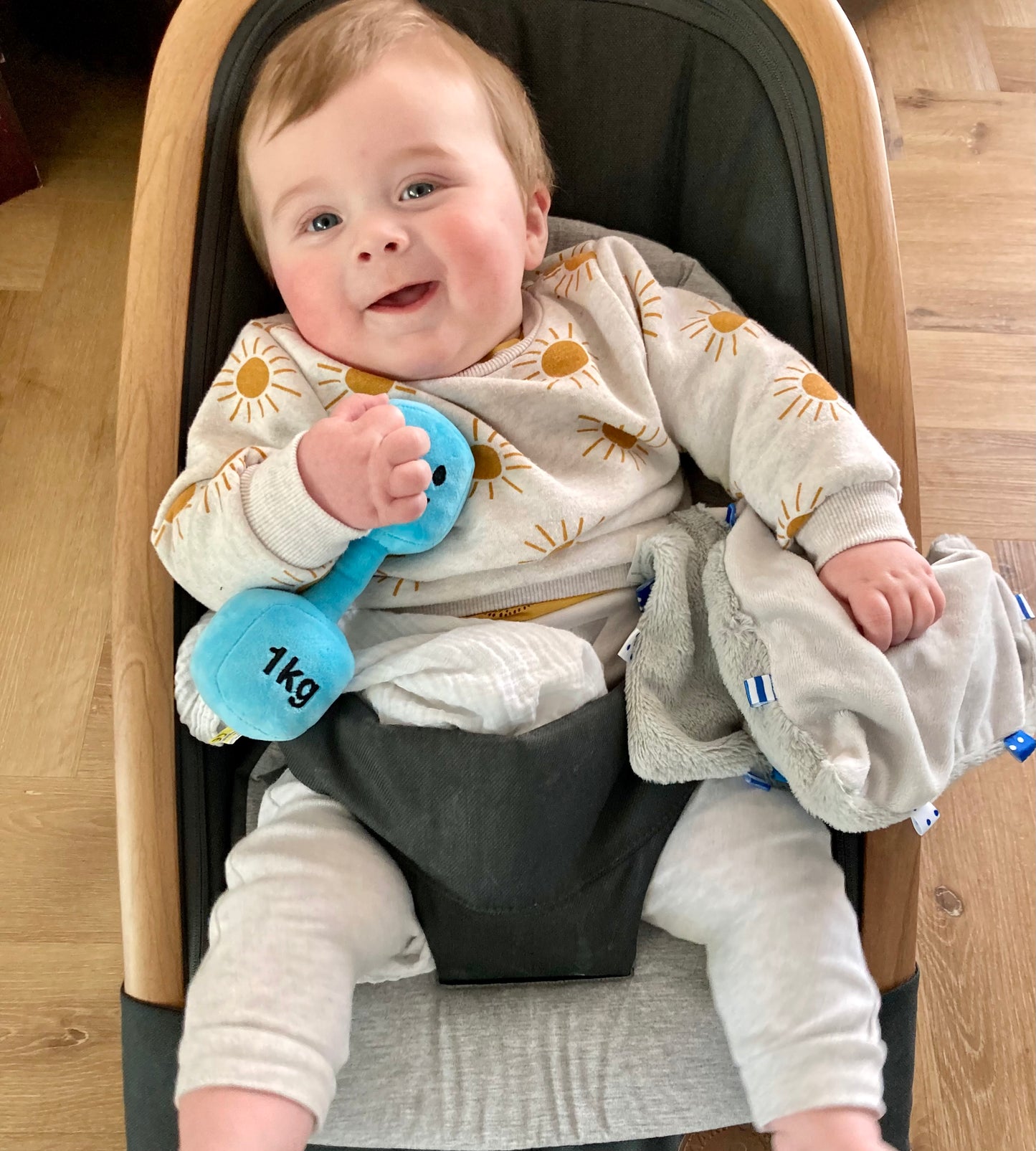 Baby in car seat with a cheeky smile, holding Hazza Toyz Mystic Blue dumbbell, enjoying playful sensory interaction during travel