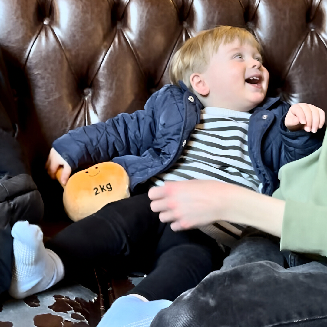 Baby laughing on Chesterfield sofa while holding Hazza Toyz Apricot Orange kettlebell, enjoying playful interaction and sensory engagement