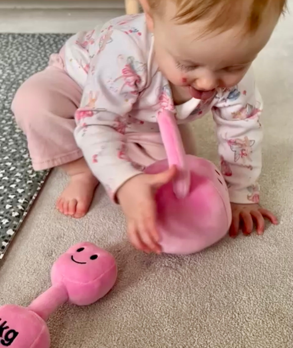 Baby crawling toward Hazza Toyz Candy Chateau Pink kettlebell with matching dumbbell in background, engaging in sensory play and motor skill development