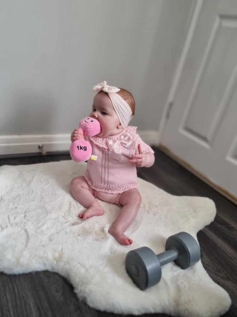 Baby girl on fluffy white rug holding and biting Hazza Toyz Candy Chateau Pink dumbbell, enjoying playful sensory exploration