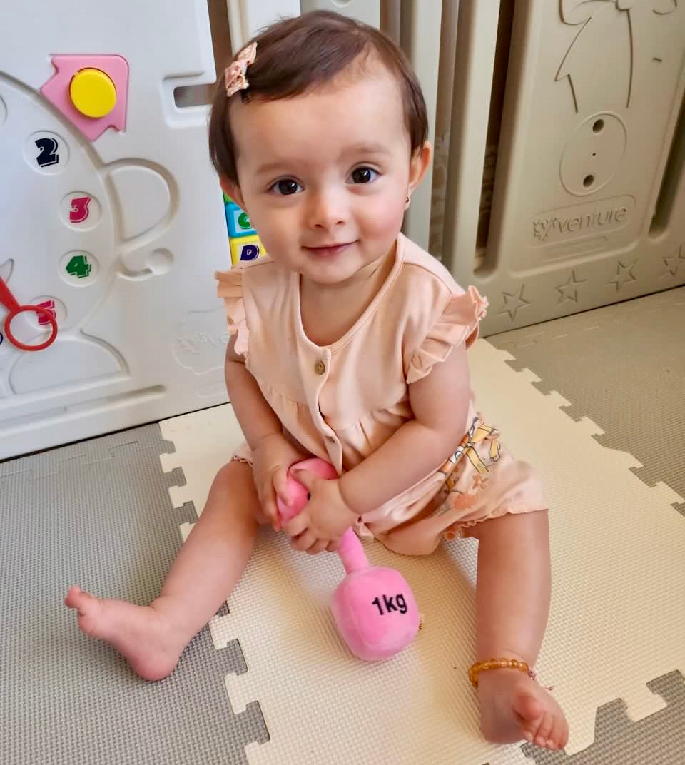 Baby girl holding Hazza Toyz Candy Chateau Pink dumbbell, enjoying playful interaction and developing motor skills