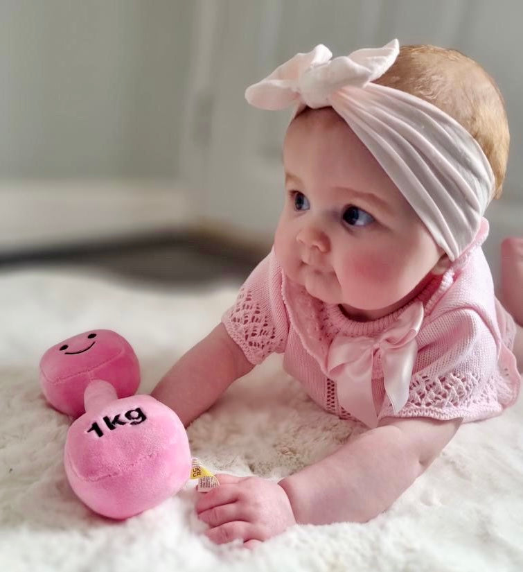 Baby girl lying on her belly on a white fluffy rug, holding Hazza Toyz Candy Chateau Pink dumbbell, enjoying soft sensory play