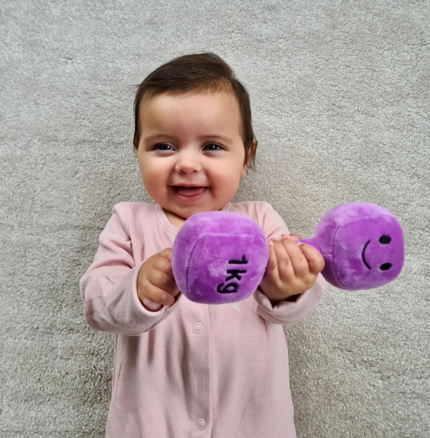 Smiling baby gripping Hazza Toyz Lavender Purple dumbbell, engaging in playful sensory development and motor skills improvement