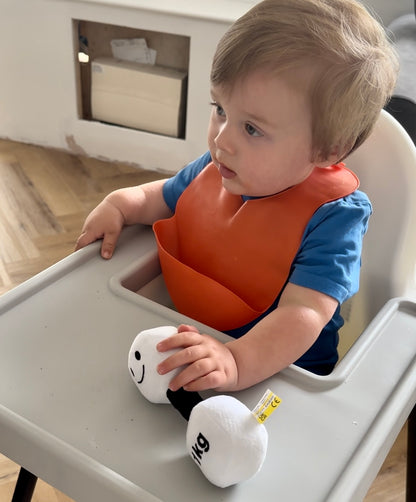 Baby in highchair wearing a bib, holding Hazza Toyz Lamby Black and White dumbbell, enjoying playful sensory engagement