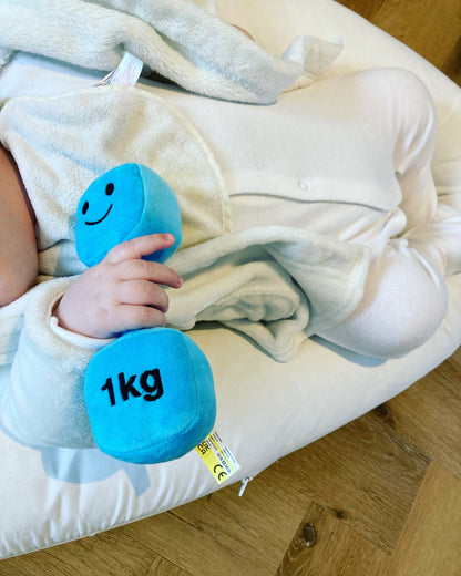 Baby in white clothes holding a blue dumbbell, enjoying playful moments with Hazza Toyz fitness-inspired toys for toddlers.