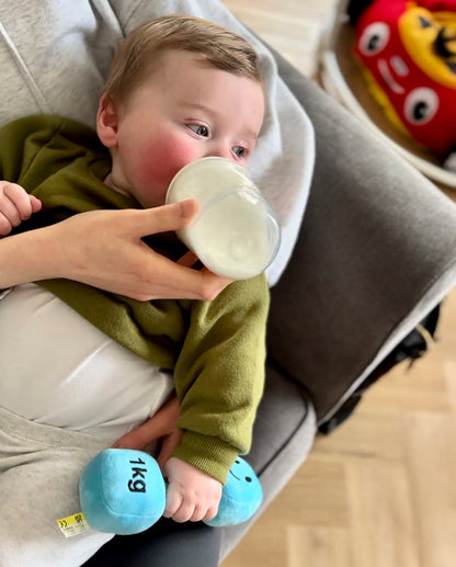 Baby on mum's lap drinking milk and holding Hazza Toyz Mystic Blue dumbbell, enjoying a comforting and playful moment