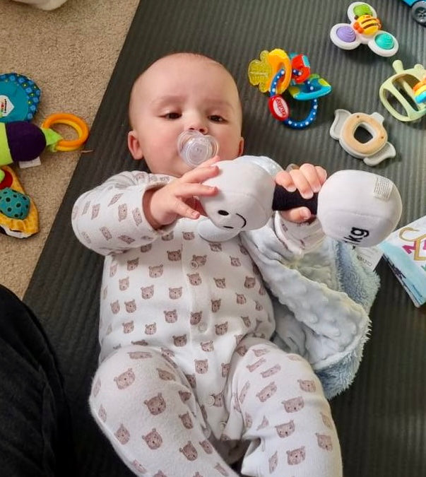 Baby on playmat surrounded by toys, holding Hazza Toyz Lamby Black and White dumbbell, engaging in sensory play and motor skill development